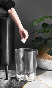 Modern transparent plastic waste bin styled next to a sofa with home decor plants