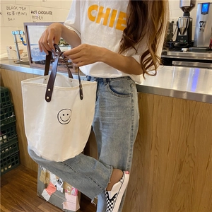 Detail of white canvas bucket bag with black smiley face and brown leather handles