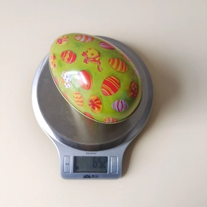 Easter egg-shaped tin box weighing 65.2 grams on a scale with colorful festive patterns
