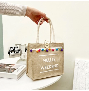 Detail of jute tote bag with 'HELLO WEEKEND' print, colorful pom poms and wooden button