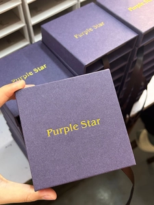 Detalle de caja de regalo cuadrada púrpura para joyas con logo dorado y lazo de cinta