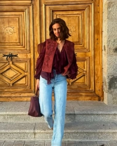 Detail of embroidered burgundy vest styled with a ruffled shirt and blue jeans in outdoor setting