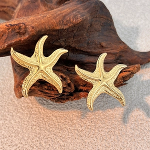 Orecchini a perno a forma di stella marina dorata con texture metallica su legno e sabbia