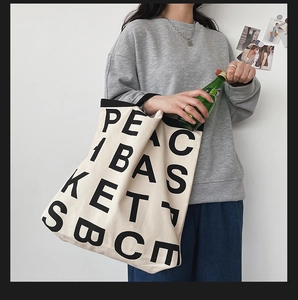 Detail of a beige canvas tote bag with bold black letter print held by a model in casual wear