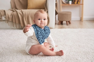 Baby wearing a blue leopard print cotton bib on a soft rug in a cozy home setting