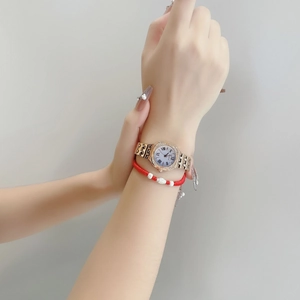 Close-up of a gold square watch with crystal bezel and a red beaded bracelet on wrist