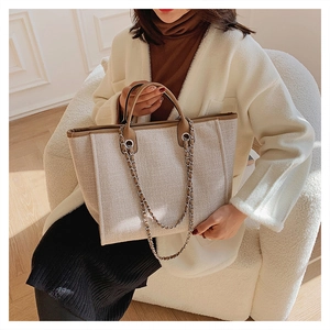 Beige woven canvas tote bag with leather handles and chain strap shown in a lifestyle setting