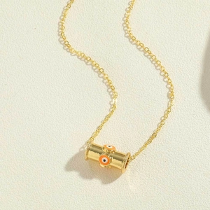 Close-up of golden cylindrical pendant with orange evil eye patterns on a fine chain