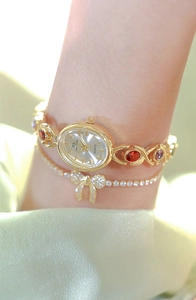 Close-up of gold oval watch with pink and red gemstones worn with a bow bracelet