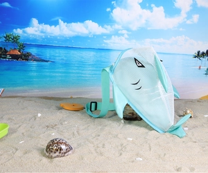 Blue shark mesh beach bag with adjustable strap on sand featuring shells and ocean view