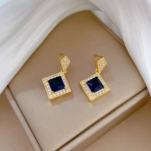 Macro shot of gold earrings with dark blue square gemstone and zircon halo on white silk