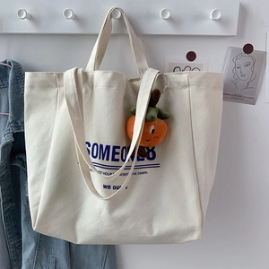 Close-up of cream canvas tote bag with blue artistic lettering and orange plush charm