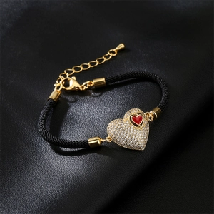 Detail of black woven rope bracelet featuring a gold heart charm with red oil drip and zircons