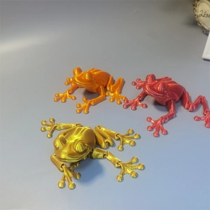 Three 3D printed articulated frogs in orange, red, and gold colors made of PLA plastic