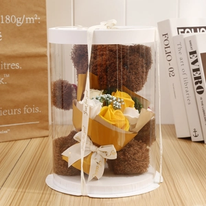 Brown teddy bear with yellow soap flowers in a clear cylindrical gift box with ribbon
