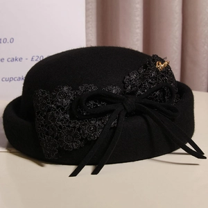 Detail of black wool beret featuring elegant floral lace, bow and gold metal logo accent