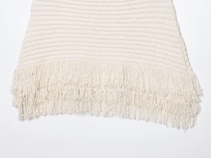 Close-up detail of cream knit fabric texture with long fringe tassels at the bottom hem