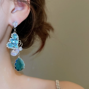 Detail of luxury silver needle earrings featuring blue butterfly and green teardrop crystals
