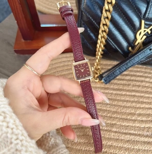 Detail of a square gold watch with burgundy leather strap held against a black quilted bag