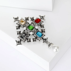 Baroque cross brooch with colorful gemstones and hanging pearl on a white pedestal