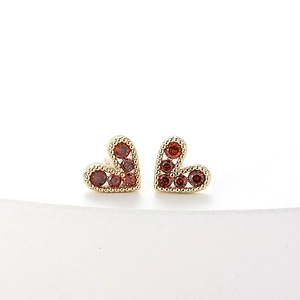 Detail of heart-shaped gold stud earrings featuring red gemstones and beaded edges
