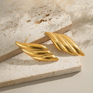 Close-up of golden leaf-shaped stainless steel earrings on a beige stone and feather background