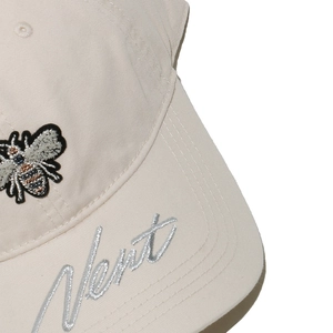 Close-up of beige baseball cap featuring bee embroidery and silver cursive Next lettering
