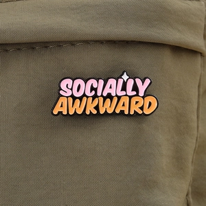 Close-up of Socially Awkward enamel pin on olive green fabric showing metal texture