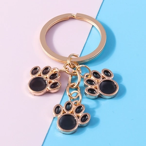 Close-up of golden keychain with three black enamel cat paw charms on pink and blue background
