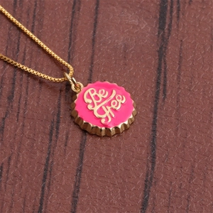 Macro detail of 18K gold-plated bottle cap pendant with Be Free script on pink enamel