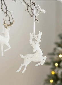 Close-up of white glittery reindeer ornament hanging on a snowy branch with warm lights