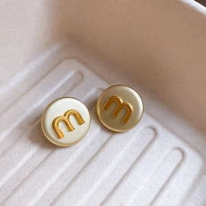 Macro shot of circular gold letter M stud earrings on a textured cream tray
