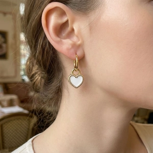 Close-up of golden heart earrings with white enamel finish worn by a model in soft light