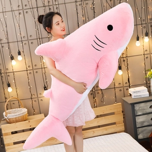Large pink shark plush toy held by a model in a cozy, warm decorated bedroom