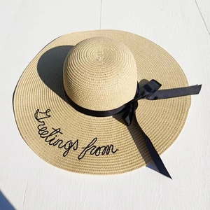 Close-up of Greetings from embroidered text on a beige wide-brim straw beach hat