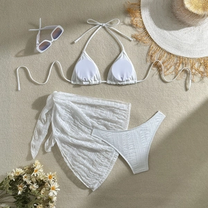 Flat lay of white three-piece bikini set with sheer mesh skirt on beach textured background