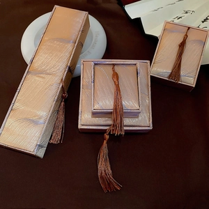 Three champagne satin jewelry boxes with tassel charms on a minimalist display stand