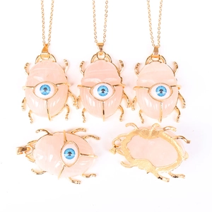 Pink gemstone beetle pendant with gold metal frame and blue eye detail on white background