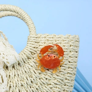 Handmade orange acrylic crab brooch accessory displayed on a beige woven summer beach bag