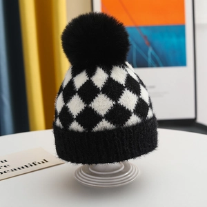 Detail of checkered knit baby hat with a large faux fur pom-pom on a white stand
