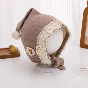 Detail of baby knit hat featuring white lace trim, flower embroidery and plush pom-poms