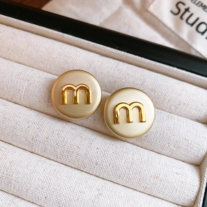 Macro detail of round cream stud earrings featuring golden letter m in display box