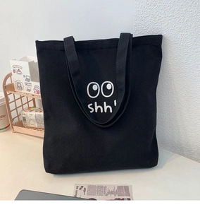 Detalle de bolso tote de lona negra con motivo de ojos y texto en mesa blanca