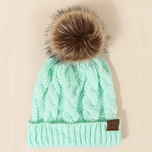 Detail of mint green cable knit beanie featuring a brown faux fur pom and a leather patch