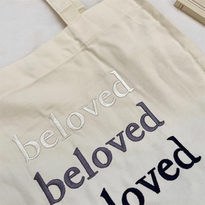 Detailed close-up of gradient beloved letter embroidery on off-white canvas tote bag