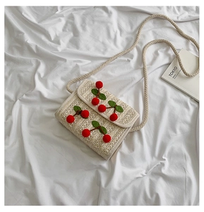 Detailed macro of beige straw crossbody bag featuring red cherry pom-poms and cotton rope strap