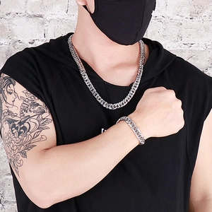 Male model wearing silver Cuban link necklace and bracelet set against white brick wall