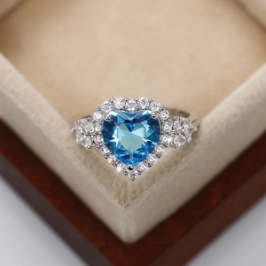Close-up of a heart-shaped blue topaz ring with diamond accents in a beige velvet box