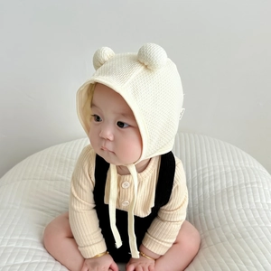 Detail of cream waffle knit baby hat with bear ears and chin straps on a soft cushion