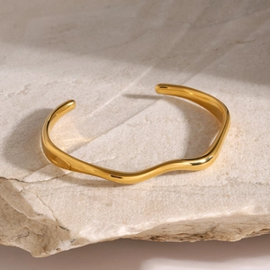Detail view of wavy gold-plated stainless steel cuff bracelet on a natural stone surface